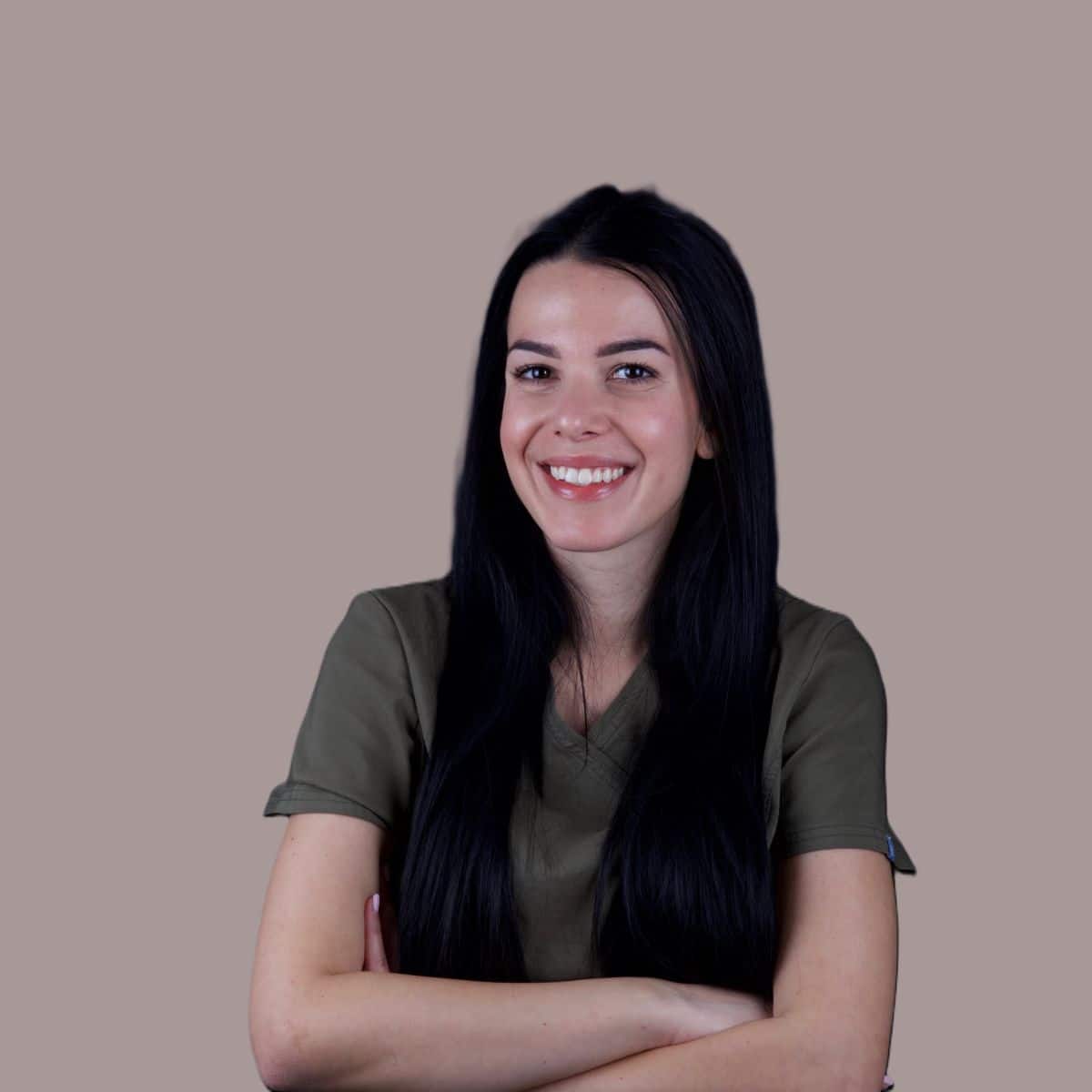 Sofija Simeunovic – White Clinic Belgrad Eine lächelnde Frau mit langen dunklen Haaren, die Arme verschränkt, in olivgrüner OP-Kleidung, Porträt vor einem taupefarbenen Hintergrund.