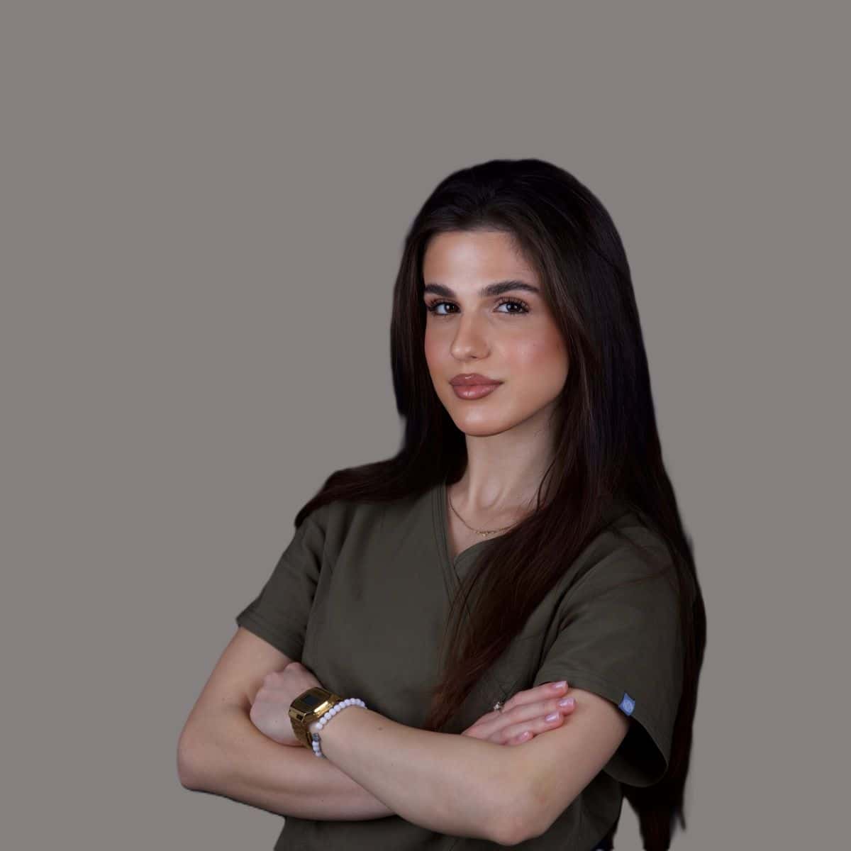 Tijana Andrejevic – White Clinic Belgrad Eine Frau mit langen dunklen Haaren, die die Arme verschränkt hat und ein olivgrünes Oberteil trägt, vor einem grauen Hintergrund.