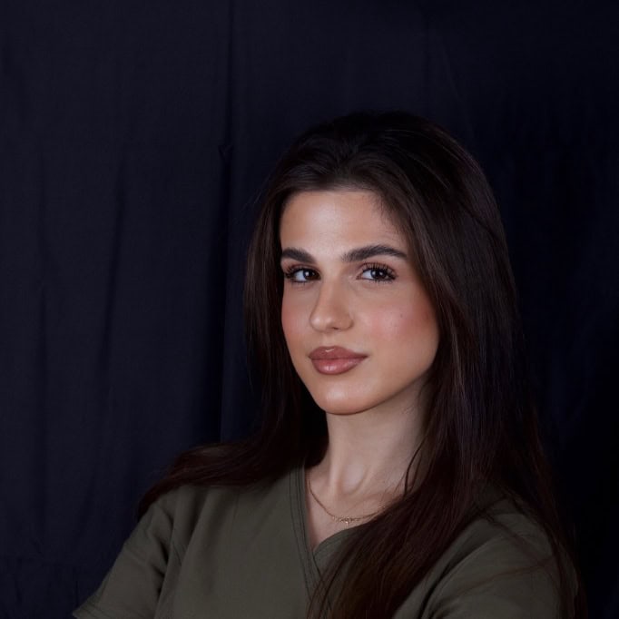 Portrait of a young woman with long dark hair and neutral makeup, looking at the camera against a dark backdrop, wearing an olive green top.