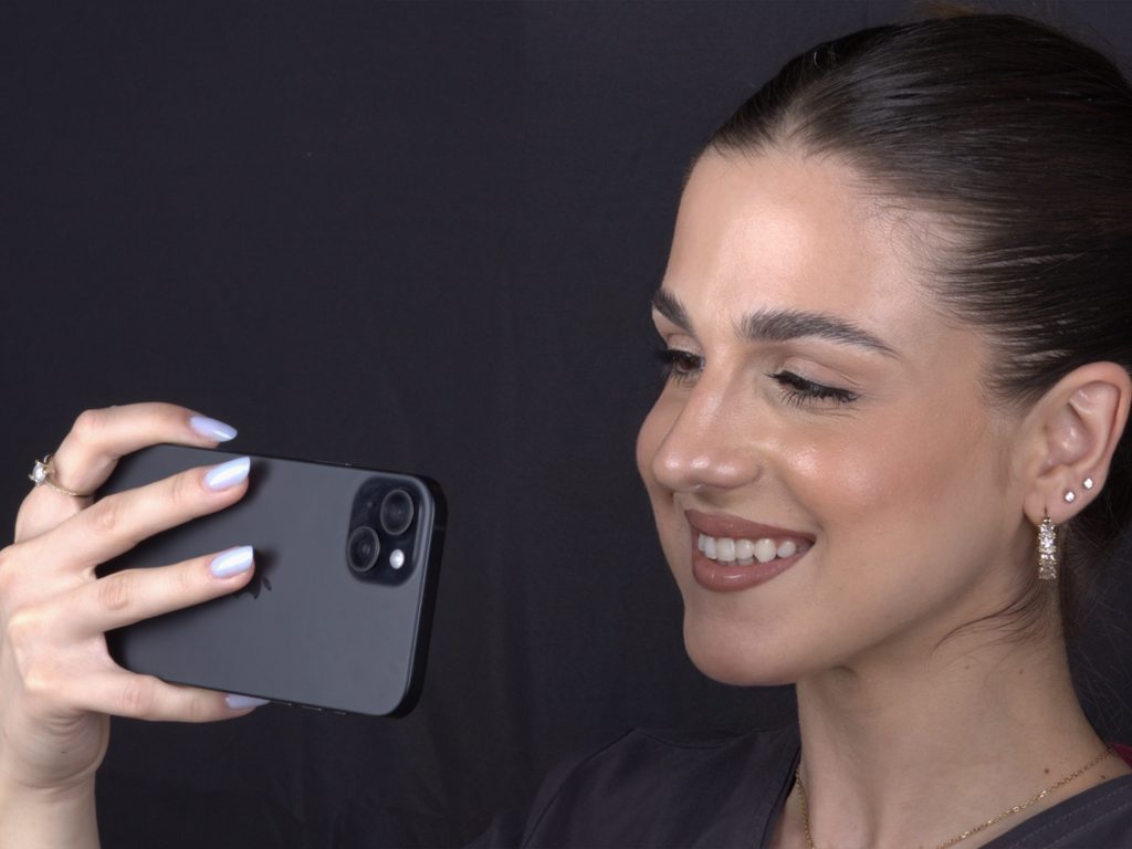 Selfie of the maximum smile - White Clinic Belgrade Woman smiling as she holds a dark smartphone to take a selfie against a black background, close up of her face and hand.