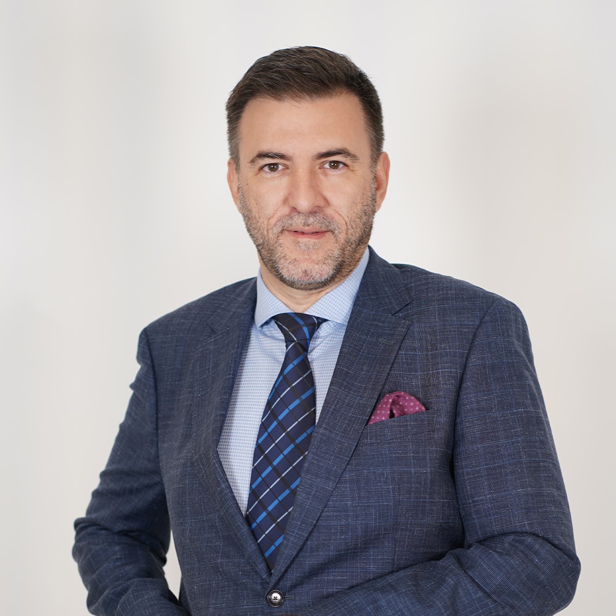 Professional headshot of a man in a navy plaid suit, light blue shirt, and blue plaid tie against a light background.