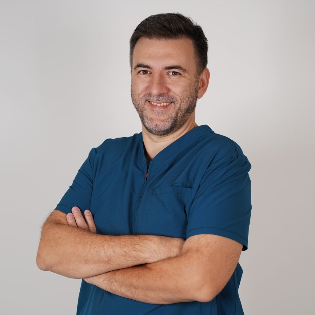 Smiling man in blue medical scrubs with arms crossed, posing against a light gray background