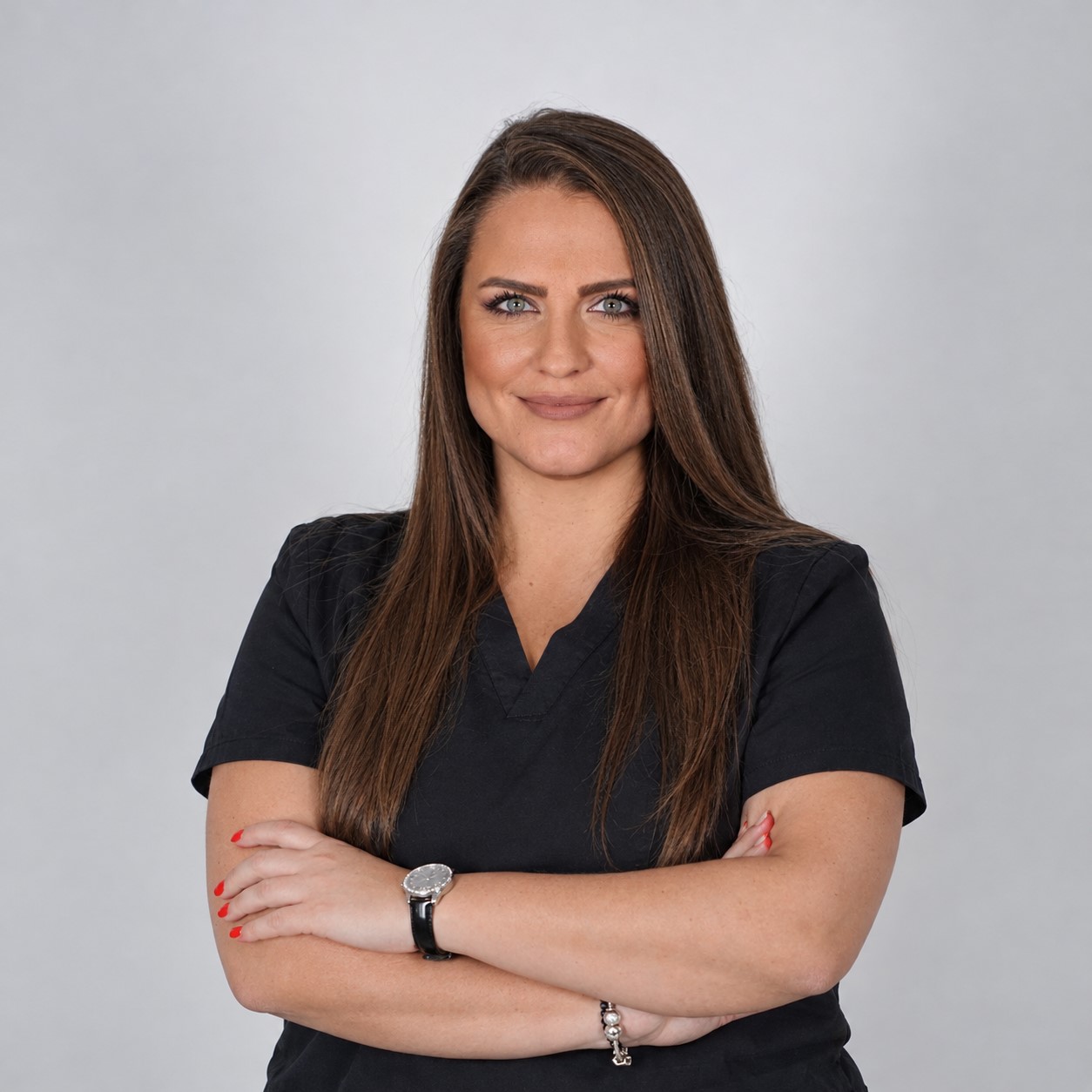 Confident professional woman with long brown hair in a black scrub top, arms crossed against a light gray background.