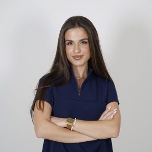 Woman with long dark hair, arms crossed, wearing an olive green top against a gray backdrop.